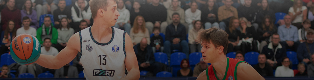 Игрок в белой форме с баскетбольным мячом противостоит игроку в зелёной форме. На фоне сидят зрители