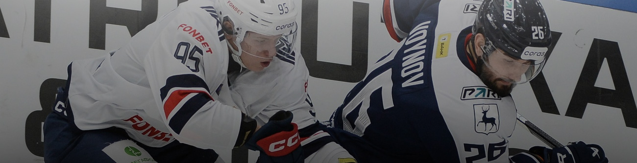 Двое хоккеистов в игровом моменте возле борта. Один игрок в белой форме, другой в тёмной. Напряжённая борьба за шайбу, близкий контакт. Спортивное действие на фоне рекламного баннера